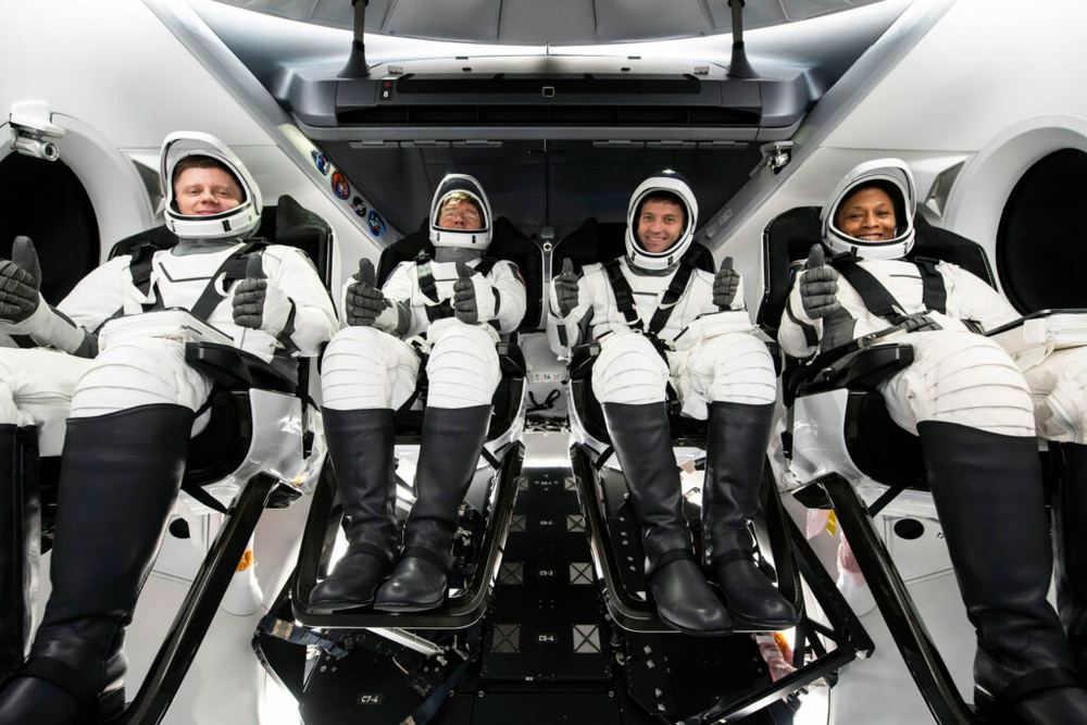 An image of four people in a capsule sitting down. They are all wearing white spacesuits. 